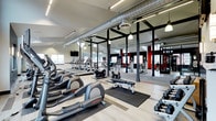 This fitness center offers a spacious, single-floor layout designed to accommodate a wide range of workout needs. The space features an industrial aesthetic, with exposed silver ductwork running across the ceiling and bold black structural columns that divide the floor into distinct workout zones while maintaining an open, airy feel. Light wood-toned flooring contrasts with the darker columns and equipment, contributing to a clean and modern atmosphere.

The cardio zone is generously stocked with elliptical trainers, upright bikes, and a row of treadmills equipped with personal screens, all positioned to take advantage of the natural light streaming through large windows. Full-length mirrors line the walls, enhancing the sense of depth and space throughout.

A dedicated free weights area includes a dumbbell rack and adjustable benches, while a separate strength and functional training zone is visible toward the back, accented by red wall panels that add a dynamic visual element. Pendant lighting supplements the natural light, ensuring the space is well-lit throughout. The overall atmosphere is energetic yet functional, making it well-suited for both focused training sessions and general fitness routines.