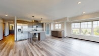 This single-floor residence is designed with a contemporary aesthetic and a functional open-concept layout. The kitchen serves as the centerpiece of the main living area, outfitted with dark espresso cabinetry, granite countertops, stainless steel appliances including a French door refrigerator, range, microwave, and dishwasher, and a central island with pendant lighting overhead. Light wood-look luxury vinyl plank flooring extends throughout the main living and kitchen areas, contributing to a bright, cohesive atmosphere. Multiple windows along the living area walls allow generous natural light to fill the space and offer views of the surrounding neighborhood. A sliding glass door opens onto a private balcony enclosed by a metal railing, providing pleasant outdoor views of green space and residential scenery. The bedrooms feature neutral carpet, large windows with natural views, and walk-in closets with wire shelving systems. The bathroom includes a tub-shower combination, a granite-topped vanity with dark cabinetry, a large mirror, and wood-look tile flooring. A dedicated laundry room with side-by-side full-size washer and dryer units and overhead shelving adds practical convenience to the overall layout.