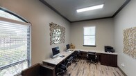 Located in Frisco, this single-story business center is thoughtfully designed to provide a functional and polished workspace. The room features built-in granite countertop workstations that wrap along the walls, offering ample surface area for multiple users. Several iMac computers are stationed along the desk, making this an ideal space for residents or visitors needing access to technology and productivity tools. Ergonomic black leather office chairs are paired with each workstation, ensuring comfort during extended use. The flooring consists of wood-look planks in a light, varied finish that adds warmth and a contemporary feel to the space. Neutral taupe walls are accented by dark crown molding, lending a refined, professional aesthetic. A decorative geometric mirror wall art piece serves as a focal point, adding visual interest and a modern touch. Natural light filters in through a window fitted with white horizontal blinds, complementing the overhead flush-mount lighting panel that ensures the space remains bright. Dark wood cabinetry beneath the countertops provides additional storage. Overall, the space balances practicality with stylish design elements.