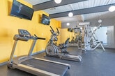 Located in Costa Mesa, California, this fitness center offers a well-equipped exercise space designed to support a full range of workout routines. The room features a striking mid-century modern aesthetic, characterized by a white-paneled ceiling with bold dark exposed beams and globe pendant lights that provide warm, even illumination throughout. Vivid yellow accent walls energize the space and are complemented by full-length mirrors along one wall, which enhance the sense of openness and allow exercisers to monitor their form. The durable rubber flooring provides a safe, comfortable surface for all types of activity. Cardio equipment includes treadmills, an elliptical trainer, and a recumbent stationary bike, all positioned beneath wall-mounted flat-screen televisions for entertainment during workouts. A multi-station cable weight machine occupies a dedicated alcove, while a dumbbell rack and stability balls round out the strength and flexibility offerings. A separate open area with a bench offers space for stretching or functional training. Large sliding glass doors along one wall invite natural light and provide views of an outdoor area, connecting the interior to the exterior environment. This fitness center is a functional and visually appealing amenity for residents at 2000 Parsons St.