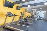 Located in Costa Mesa, California, this fitness center offers a well-equipped exercise space designed to support a full range of workout routines. The room features a striking mid-century modern aesthetic, characterized by a white-paneled ceiling with bold dark exposed beams and globe pendant lights that provide warm, even illumination throughout. Vivid yellow accent walls energize the space and are complemented by full-length mirrors along one wall, which enhance the sense of openness and allow exercisers to monitor their form. The durable rubber flooring provides a safe, comfortable surface for all types of activity. Cardio equipment includes treadmills, an elliptical trainer, and a recumbent stationary bike, all positioned beneath wall-mounted flat-screen televisions for entertainment during workouts. A multi-station cable weight machine occupies a dedicated alcove, while a dumbbell rack and stability balls round out the strength and flexibility offerings. A separate open area with a bench offers space for stretching or functional training. Large sliding glass doors along one wall invite natural light and provide views of an outdoor area, connecting the interior to the exterior environment. This fitness center is a functional and visually appealing amenity for residents at 2000 Parsons St.