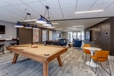 This contemporary amenity lounge is thoughtfully designed to accommodate both active recreation and relaxed socializing. The space is unified by patterned carpet flooring, recessed linear LED lighting, and a sophisticated color palette of charcoal, grey, and white, punctuated by bold yellow and blue accent furnishings.

The game area anchors one end of the space, featuring a full-size pool table illuminated by a trio of hanging pendant lights, alongside a foosball table. A large wall-mounted flat-screen television is positioned above a seating cluster of plush blue armchairs arranged around a round coffee table — ideal for media viewing or casual conversation.

Adjacent to the game area, a dedicated lounge zone showcases a curated gallery wall of vibrant abstract artwork displayed against a dark charcoal accent wall, creating a striking focal point. Comfortable blue accent chairs with decorative pillows are arranged in conversational groupings throughout.

Bistro-style seating with marble-top round tables and bright yellow chairs adds a lively, social dimension to the space. A hallway corridor featuring textured wallcovering and framed photography connects the areas seamlessly. Glass partitions offer views into adjacent spaces, including a stairwell, enhancing the open and connected feel. The overall ambiance is modern, energetic, and inviting.