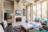 This elegant single-floor space serves as a refined communal or leasing hub, thoughtfully designed with upscale architectural details throughout. The grand entrance foyer greets visitors with soaring ceilings, a statement chandelier, rich hardwood-style French doors, large transom windows, and light tile flooring that flows seamlessly throughout the space.

The main lounge area is a standout, boasting an impressive coffered ceiling with ornate crown molding and an abundance of floor-to-ceiling windows that flood the interior with natural light and offer views of a lush outdoor pool area. A classic white-mantled fireplace with a brick surround serves as a focal point, complemented by a wall-mounted flat-screen TV above. Stylish patterned area rugs, contemporary accent chairs with teal throw pillows, and decorative artwork contribute to the space's polished, welcoming ambiance.

An adjacent open seating area features dramatic double-height ceilings with recessed lighting, additional lounge seating, and direct sightlines to a kitchen bar area with counter seating—ideal for meetings or casual gatherings. The kitchen itself is well-appointed with white cabinetry, dark mosaic tile backsplash, rich countertops, a stainless steel double sink, and a dishwasher, all brightened by a pair of windows overlooking a brick exterior courtyard.