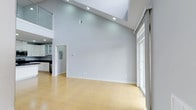 This two-story, 2-bedroom, 2-bathroom residence offers a thoughtfully designed layout that maximizes both space and comfort. The main living area immediately impresses with its soaring vaulted ceilings, creating an open and airy atmosphere that feels expansive and welcoming. Recessed lighting is strategically placed throughout, complementing the generous natural light that pours in through large sliding glass doors and windows, seamlessly connecting the interior to the outdoors. The light-toned hardwood flooring adds warmth to the cool, neutral palette of the walls, lending the space a clean and modern aesthetic. The kitchen is visible from the main living area and features dark cabinetry contrasted against glass-front upper cabinets, offering both style and functionality. A second-floor loft or upper level overlooks the main living space, adding to the dramatic vertical scale and providing additional versatility. The open-concept flow between the living area and kitchen makes this residence well-suited for both everyday living and entertaining. The overall ambiance is bright, contemporary, and inviting, with a minimalist design that allows the architectural details — particularly the striking ceiling height — to take center stage.