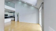 This two-story, 2-bedroom, 2-bathroom residence offers a thoughtfully designed layout that maximizes both space and comfort. The main living area immediately impresses with its soaring vaulted ceilings, creating an open and airy atmosphere that feels expansive and welcoming. Recessed lighting is strategically placed throughout, complementing the generous natural light that pours in through large sliding glass doors and windows, seamlessly connecting the interior to the outdoors. The light-toned hardwood flooring adds warmth to the cool, neutral palette of the walls, lending the space a clean and modern aesthetic. The kitchen is visible from the main living area and features dark cabinetry contrasted against glass-front upper cabinets, offering both style and functionality. A second-floor loft or upper level overlooks the main living space, adding to the dramatic vertical scale and providing additional versatility. The open-concept flow between the living area and kitchen makes this residence well-suited for both everyday living and entertaining. The overall ambiance is bright, contemporary, and inviting, with a minimalist design that allows the architectural details — particularly the striking ceiling height — to take center stage.
