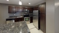 This single-floor residence showcases a thoughtfully designed layout that balances functionality and modern style throughout. The kitchen serves as a standout feature, boasting rich dark-stained cabinetry, granite countertops with subtle veining, and a full suite of stainless steel appliances including a refrigerator, range, and built-in microwave. Large-format white tile flooring with inlaid accent lines runs through the kitchen and extends into the hallways, lending a clean, polished aesthetic. A generous peninsula provides additional counter space and casual seating potential. The living areas transition to plush wall-to-wall carpeting, offering a warm and comfortable feel underfoot. Bedrooms are well-sized, each benefiting from natural light through windows with exterior views, and neutral gray wall tones create a versatile backdrop. Storage is a priority, with at least one expansive walk-in closet featuring built-in wire shelving along multiple walls. The bathroom is finished with the same cohesive design language — dark cabinetry, granite vanity top, a large mirror with lighted bar, and a tub/shower combination set within a bright, tiled surround. Recessed and pendant lighting throughout ensures well-lit spaces at every turn.