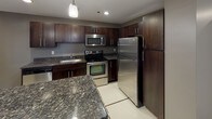 This single-floor residence showcases a thoughtfully designed layout that balances functionality and modern style throughout. The kitchen serves as a standout feature, boasting rich dark-stained cabinetry, granite countertops with subtle veining, and a full suite of stainless steel appliances including a refrigerator, range, and built-in microwave. Large-format white tile flooring with inlaid accent lines runs through the kitchen and extends into the hallways, lending a clean, polished aesthetic. A generous peninsula provides additional counter space and casual seating potential. The living areas transition to plush wall-to-wall carpeting, offering a warm and comfortable feel underfoot. Bedrooms are well-sized, each benefiting from natural light through windows with exterior views, and neutral gray wall tones create a versatile backdrop. Storage is a priority, with at least one expansive walk-in closet featuring built-in wire shelving along multiple walls. The bathroom is finished with the same cohesive design language — dark cabinetry, granite vanity top, a large mirror with lighted bar, and a tub/shower combination set within a bright, tiled surround. Recessed and pendant lighting throughout ensures well-lit spaces at every turn.
