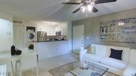 This single-floor space offers a clean, open-concept layout that seamlessly connects the living and kitchen areas, creating a sense of spaciousness and easy flow throughout. The living area is bright and welcoming, featuring light-toned tile flooring, a ceiling fan with integrated lighting, and a cozy seating arrangement centered around a glass-top coffee table. Neutral gray and white walls provide a fresh, modern backdrop complemented by tasteful wall art.

The kitchen is designed for both functionality and style, featuring white cabinetry, dark countertops, and stainless steel appliances including a refrigerator and microwave. A raised breakfast bar divides the kitchen from the living space, offering additional seating and an interactive cooking environment. Track lighting above the kitchen ensures ample task illumination.

A notable convenience of this space is the in-unit stacked washer and dryer, positioned just off the kitchen area, adding practical day-to-day functionality. A white interior door leads to additional areas of the unit, maintaining a cohesive, clean aesthetic throughout. The overall ambiance is light, airy, and modern, making it a well-appointed and efficiently organized living space.