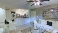 This single-floor space offers a clean, open-concept layout that seamlessly connects the living and kitchen areas, creating a sense of spaciousness and easy flow throughout. The living area is bright and welcoming, featuring light-toned tile flooring, a ceiling fan with integrated lighting, and a cozy seating arrangement centered around a glass-top coffee table. Neutral gray and white walls provide a fresh, modern backdrop complemented by tasteful wall art.

The kitchen is designed for both functionality and style, featuring white cabinetry, dark countertops, and stainless steel appliances including a refrigerator and microwave. A raised breakfast bar divides the kitchen from the living space, offering additional seating and an interactive cooking environment. Track lighting above the kitchen ensures ample task illumination.

A notable convenience of this space is the in-unit stacked washer and dryer, positioned just off the kitchen area, adding practical day-to-day functionality. A white interior door leads to additional areas of the unit, maintaining a cohesive, clean aesthetic throughout. The overall ambiance is light, airy, and modern, making it a well-appointed and efficiently organized living space.
