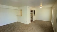 This single-floor residential unit offers a straightforward and functional layout designed for everyday living. The main living area is finished with wall-to-wall carpet in a neutral tone, providing a comfortable and cohesive foundation throughout the space. Walls are painted in a clean, light off-white, contributing to a bright and open atmosphere.

A defining architectural feature is the partial dividing wall that separates the living area from the kitchen. This wall incorporates a pass-through counter with a built-in shelf, allowing for convenient interaction between the two spaces while maintaining a degree of visual separation. The pass-through design enhances the sense of openness and connectivity within the unit.

The kitchen is fully equipped with standard white appliances, including a range with oven and an overhead microwave or range hood. Dark upper cabinetry provides storage and a subtle contrast to the otherwise light interior palette. The kitchen also features its own pass-through counter area, reinforcing the open-concept feel.

Overhead lighting is provided via a flush-mount ceiling fixture in the living area, ensuring adequate illumination. A ceiling vent is also visible, indicating a central HVAC system for climate control. Overall, the space presents a clean, move-in ready environment with a practical and efficient design.