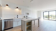 This single-floor residence showcases a clean, contemporary design with a bright and airy open-concept layout. Light-toned hardwood flooring runs throughout the main living areas, creating a warm and cohesive aesthetic.

The kitchen is a standout feature, boasting white shaker-style cabinetry, a vertical tile backsplash, a farmhouse sink with a matte black faucet, and stainless steel appliances including a refrigerator, electric range, and dishwasher. A central island with built-in shelving provides additional workspace and storage. Pendant lighting with copper-toned fixtures adds a stylish accent above the kitchen area.

The open living space flows seamlessly from the kitchen and is bathed in natural light, with large black-framed sliding glass doors leading to a balcony with outdoor views of surrounding greenery. The bedrooms feature plush neutral carpeting and floor-to-ceiling windows that invite abundant daylight and pleasant exterior views.

The bathroom is well-designed with white cabinetry, a wide vanity countertop, matte black hardware, and a large frameless mirror. A separate toilet area and wood-look flooring add both function and style. Throughout the residence, the consistent use of white walls, black hardware accents, and natural materials creates a cohesive modern aesthetic.