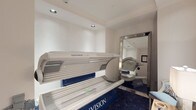 This tanning studio room is a compact, well-appointed space designed to deliver a professional and comfortable tanning experience. At its centerpiece sits a SunVision Elite tanning bed, a high-end unit with a distinctive blue and white finish, positioned prominently within the room. The flooring features a striking deep blue carpet with a geometric chevron-style pattern, lending the space a polished, boutique aesthetic that contrasts elegantly with the clean white walls and recessed ceiling lighting above.

A large, full-length floor mirror with a sleek chrome-framed border adds both a functional and decorative element, reflecting the room and creating a sense of added depth. A cushioned accent chair with a light blue seat and patterned throw pillow provides a comfortable spot for clients before or after their session. A small wooden side table holds tanning lotions, accessories, and a softly lit table lamp, enhancing the welcoming ambiance of the room.

The entry door is clean white with standard hardware, and a framed informational notice is mounted nearby on the wall, consistent with professional tanning facility standards. The overall design strikes a balance between functionality and refined comfort, making this studio room feel both approachable and upscale — a purpose-built environment for an efficient and enjoyable tanning experience.