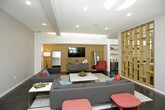 This thoughtfully designed single-floor communal amenity space blends contemporary style with practical functionality across several distinct yet interconnected zones. The open-plan layout flows seamlessly between areas, unified by polished dark concrete flooring, recessed lighting, and a neutral gray and white color palette accented by warm wood tones and bold pops of teal, coral, and chartreuse.

The main lounge area is anchored by a wood-paneled feature wall with a mounted flat-screen TV, complemented by a sectional sofa, coordinating armchairs, and vivid accent ottomans arranged around a marble-topped coffee table. An open-shelved golden room divider elegantly separates this zone from the rest of the space while maintaining visual connectivity throughout.

The communal kitchen is a standout feature, outfitted with dark wood cabinetry, white quartz countertops, a gas cooktop island with bar seating, a stainless steel range hood, white subway tile backsplash, and open yellow floating shelves for display and storage. A beverage station is conveniently located nearby.

Adjacent to the kitchen, a casual dining and lounge area offers high-top tables with mixed seating and upholstered booth-style banquettes, bathed in natural light from large windows. Glass doors lead directly to an outdoor deck, further extending the usable social space and bringing in abundant daylight.