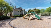 This outdoor playground is a well-appointed recreational amenity situated within a residential community. The centerpiece is a multi-level play structure finished in green and tan, featuring multiple slides, enclosed tunnel sections, and elevated platforms designed to engage children of various ages. The structure is set on a generous gravel surface, which provides impact-absorbing ground cover across the play area.

The surrounding environment is thoughtfully landscaped, with mature trees, neatly trimmed shrubs, and maintained green spaces that contribute to a pleasant, park-like atmosphere. Curved concrete pathways wind through the grounds, lined with crape myrtle trees and mulched beds that add visual appeal throughout the seasons.

Adjacent to the playground, a covered entrance area features classic lap siding, decorative lighting, and black metal railing details consistent with the broader community's architectural character. The buildings nearby reflect a traditional residential design with pitched rooflines and neutral exterior finishes that blend naturally into the tree-lined setting.

The open-air setting benefits from abundant natural light and open sky views, creating a bright and inviting environment. An emergency phone station is also present near the entrance, adding a practical safety feature to the space. Overall, the playground offers a functional and attractive outdoor destination within a well-maintained community.