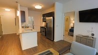 This single-floor residence offers a thoughtfully designed open-concept layout that seamlessly connects the kitchen, dining, and living areas. The kitchen is a standout feature, equipped with stainless steel appliances including a French door refrigerator, a gas range with microwave above, and dark espresso cabinetry that provides ample storage. A kitchen island with a granite countertop and an undermount sink adds both functionality and style, serving as a natural dividing point between the kitchen and the living space. Rich hardwood-style flooring extends throughout the main living area, adding warmth and visual continuity. Flush-mount ceiling lighting provides even, ambient illumination throughout the space. The living area flows naturally from the kitchen, offering a comfortable and versatile space. A doorway visible from the main area leads into a carpeted bedroom, which features a walk-in closet and a softly lit, cozy ambiance. The white walls and clean architectural lines throughout create a bright, modern atmosphere. Overall, this residence combines practical design with contemporary finishes to deliver a comfortable and stylish living environment.