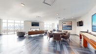 Sky Terrace is a sophisticated, single-floor amenity space offering a generous open-concept layout designed for both entertaining and relaxation. The interior is anchored by striking herringbone-patterned flooring in cool blue-gray tones, which flows seamlessly throughout the entire space and contributes to its modern, upscale aesthetic.

The fully equipped kitchen features an expansive white quartz island with bar seating, sleek white cabinetry, stainless steel appliances including a full-size refrigerator and range, and a marble-inspired tile backsplash. A series of white dome pendant lights illuminate the workspace, adding warmth and character.

The living area centers around a textured white feature wall with a built-in fireplace and a wall-mounted television, complemented by a curated mix of leather and upholstered seating. Floor-to-ceiling windows flood the space with natural light and frame breathtaking panoramic skyline views, with direct access to an outdoor terrace furnished for al fresco dining.

A dedicated game area includes a billiards table positioned beneath a statement pendant light, alongside a gaming and lounge zone with a round table and leather chairs. Curated artwork throughout adds a gallery-like quality, completing this elevated communal retreat.