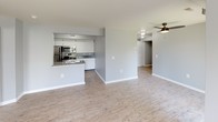 This single-floor residence features a thoughtfully designed open-concept layout connecting the living area and kitchen. The living space is finished with light wood-style flooring and neutral gray walls, with a window dressed in vertical blinds allowing natural light to filter in. A ceiling fan overhead adds both function and airflow to the space.

The kitchen is positioned adjacent to the living area, separated by a half-wall peninsula that doubles as additional counter space. White cabinetry extends to the ceiling, providing generous storage, while stainless steel appliances—including a refrigerator, range, and built-in microwave—lend a modern aesthetic. A double sink with a granite-style countertop completes the space.

The bedrooms feature plush carpeting and neutral gray walls, with windows offering views of surrounding greenery. The primary bedroom includes direct access to an en-suite bathroom and a dedicated closet. The en-suite bathroom is equipped with a walk-in shower featuring a frosted glass door, a granite-style vanity, and a decorative framed mirror. A second full bathroom mirrors this design with the addition of a soaking tub and shower combination.