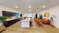 This single-floor community space is thoughtfully designed to accommodate a variety of activities and social interactions. The open-concept layout is anchored by light-toned hardwood-style flooring that flows seamlessly throughout, creating a cohesive and inviting atmosphere. Recessed lighting illuminates the interior evenly, while multiple windows along the perimeter allow natural light to brighten the space and provide views of the surrounding outdoors.

The main gathering area features a central island with a light granite-style countertop and bar-height seating, functioning as a natural focal point. Along one wall, an extended kitchenette with dark espresso cabinetry, light countertops, and a dedicated coffee bar station provides practical amenities for residents or guests. Colorful braided rugs define functional zones within the open floor plan.

The lounge area is furnished with rich cognac leather sofas and accent chairs, complemented by bold, vibrant artwork and lush potted plants that add warmth and visual interest. A secondary seating nook features blue upholstered chairs arranged around a decorative console with an ornate mirror. The floorplan also incorporates a fitness area with exercise equipment and a children's activity zone with table seating. Dual drinking fountains are accessible near the entry corridor, reflecting the space's community-oriented design.