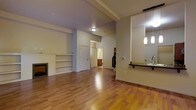 This two-floor space offers a thoughtfully designed layout that balances open living areas with functional room connections. The main living area is a standout, showcasing warm-toned hardwood flooring that extends throughout, creating a cohesive and inviting atmosphere. A built-in fireplace anchors one wall, flanked by built-in shelving that provides both display and storage opportunities. Exposed beam detailing along the ceiling adds architectural character and a sense of structure to the space.

The kitchen is integrated seamlessly into the open-concept layout, featuring a dark countertop peninsula with pendant lighting above and a large mirror that enhances the sense of depth and light in the area. A sink with a modern faucet completes the kitchen workspace. The neutral wall tones complement the natural wood flooring and contribute to the light, airy feel of the interior.

Doorways from the main living area lead to additional rooms, including what appears to be a bathroom with warm wood cabinetry visible in the background, as well as access to a staircase connecting the two floors. The overall ambiance is clean, open, and well-lit, making this a versatile and functional multi-level space suited to a variety of uses.