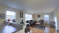 This single-floor residence offers a thoughtfully designed open-concept layout that connects the kitchen and living areas into one cohesive, welcoming space. The kitchen features a raised bar counter with seating, making it an ideal spot for casual dining or entertaining guests. Warm hardwood flooring flows throughout, adding a natural richness to the interior. The living area is generously proportioned, comfortably accommodating a full sofa and seating arrangement centered around a coffee table, all anchored by a large area rug. Multiple windows fitted with white horizontal blinds line the walls, flooding the space with natural light and creating a bright, airy atmosphere. The neutral, cream-toned walls provide a clean and versatile backdrop, complemented by tasteful wall art that adds subtle character. Ceiling height is standard and consistent throughout, contributing to the room's comfortable and uncluttered feel. The open sightlines from the kitchen counter into the living room enhance the sense of spaciousness, making this layout particularly functional for everyday living. Overall, the space presents a practical yet inviting environment with a balanced blend of casual comfort and clean, modern aesthetics.