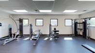 This fitness facility is designed to support a full range of workout activities within a clean, functional, and well-organized single-floor layout. The space features a drop-panel ceiling with recessed fluorescent lighting that ensures consistent, even illumination throughout. A series of square windows along the walls allow natural light to filter in, creating bright patches across the dark rubber flooring and contributing to an energizing atmosphere.

The facility is outfitted with a variety of professional-grade strength training machines, including cable and pulley systems, chest press, leg extension, and lat pulldown equipment. A dedicated free weights area includes a multi-tier dumbbell rack stocked with a wide range of weights, accompanied by adjustable workout benches for versatile training options. A wall-mounted mirror in the free weights section supports proper form and technique.

Cardio equipment, including elliptical trainers and similar machines, is visible and provides options for aerobic exercise. A wall-mounted flat-screen television adds an entertainment element to the space. Additional amenities include a water cooler station and a waste receptacle, ensuring convenience and cleanliness. The neutral-toned walls and durable flooring reflect a practical, no-frills design focused on functionality and ease of use.
