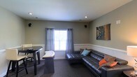 This single-floor space offers a well-appointed and inviting layout designed for both relaxation and casual gathering. The main living area is anchored by a large dark leather sectional sofa adorned with colorful accent pillows, creating a comfortable and stylish focal point. Recessed lighting in the ceiling provides warm, even illumination throughout the room, complementing the natural light that filters in through a window dressed with sheer gray curtain panels over horizontal blinds. The walls feature a neutral gray-green tone accented by white wainscoting along the lower half, lending the space a clean, classic character. A piece of abstract wall art adds a creative touch to the room's aesthetic. Adjacent to the lounge area is a compact dining setup with a round dark table and seating, ideal for casual meals or conversation. The carpeted flooring in a deep, textured pattern ties the space together while adding warmth underfoot. Ceiling-mounted speakers suggest the space is equipped for audio entertainment. Overall, the room presents a balanced combination of comfort, functionality, and understated style.