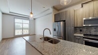 This well-appointed single-floor residence offers a thoughtfully designed open-concept layout that seamlessly connects the kitchen and living areas. The kitchen serves as the centerpiece of the space, showcasing granite countertop islands with an undermount double sink and a sleek pull-down faucet. Wood-tone cabinetry extends floor to ceiling, providing generous storage, while a mosaic tile backsplash adds a refined decorative touch. Stainless steel appliances — including a French door refrigerator with ice dispenser, a gas range, and an over-the-range microwave — complete the kitchen's modern aesthetic. Stylish glass pendant lights hang above the island and living areas, contributing to the warm, contemporary ambiance. Crown molding and tray ceiling details elevate the living area, which features warm wood-look flooring and large windows that invite abundant natural light. The bedroom offers plush carpeting, a ceiling fan, and a bright window with outdoor views, creating a comfortable retreat. An additional room with wood-look tile flooring and a tray ceiling provides flexible living space. The bathroom is finished with large-format marble-look tile flooring and a glass-enclosed walk-in shower, reflecting a clean and modern design sensibility throughout the residence.