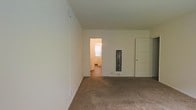 This single-floor space presents a straightforward, functional layout with clean white walls throughout. The main living area is finished with wall-to-wall carpet in a neutral tone, providing a practical and understated foundation for the room. The walls are smooth and painted white, lending a bright, open feel to the interior.

A doorway opening into an adjacent room is visible from the main living area, where natural light filters through a window, illuminating the connected space. The adjoining room appears to have hard flooring, offering a contrast to the carpeted main area. A narrow decorative window with ornamental ironwork detail is situated along one wall, adding a subtle architectural accent to the otherwise minimal interior.

A paneled door on the far wall provides access to an additional room or closet, contributing to the overall connectivity of the floor plan. A wall-mounted heating unit is visible near the baseboard, indicating an independent heating source for the space. The layout is open and uncluttered, making the space adaptable for a variety of uses. Overall, the interior maintains a clean, no-frills aesthetic with functional details throughout.