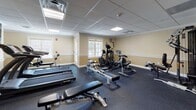 Located at 21 Dexter Rd in Foxboro, Massachusetts, this fitness center offers a well-equipped, single-floor workout facility designed to support a full range of exercise routines. The space features a drop-panel ceiling with recessed lighting that ensures consistent, even illumination throughout. Durable rubber flooring covers the entire workout area, providing safety and comfort underfoot. 

The cardio section is equipped with SportsArt treadmills, an elliptical trainer, a stationary bike, and rowing machines, giving users a variety of aerobic options. A wall-mounted television accompanies the cardio zone, and a large mirror along one wall enhances the sense of space while allowing users to monitor their form. 

For strength training, the facility includes a multi-station cable and weight machine, a dedicated dumbbell rack with a full range of free weights, an adjustable workout bench, and additional resistance equipment. Natural light filters in through several windows, contributing to an open and inviting atmosphere. 

The warm beige walls, accented with white chair rail molding and crown molding details, give the space a polished, well-maintained appearance. Additional amenities include restroom access and clear exit signage, ensuring both convenience and safety for all users.
