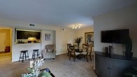 This single-floor residence offers a thoughtfully arranged open-concept layout that seamlessly connects the living, dining, and kitchen areas into one cohesive and functional space. The living area is carpeted in neutral tones, providing a warm and comfortable foundation that complements the soft, cream-colored walls throughout. A cozy accent chair adds a relaxed seating option alongside the main living furnishings, while decorative greenery and artwork contribute to the welcoming ambiance.

The dining area is centrally positioned and illuminated by a modern track lighting fixture mounted to the ceiling, casting focused light over a classic wood and glass dining table surrounded by traditional-style chairs. A large framed artwork anchors the dining space against the wall, adding a refined decorative touch.

Adjacent to the dining and living areas, the kitchen features dark cabinetry and is accessible via a convenient pass-through counter with bar stool seating, making it ideal for casual dining or entertaining. The kitchen is visibly well-equipped and efficiently laid out.

A wall-mounted flat-screen television near the living area adds a practical modern element to the space. The textured ceiling, visible ventilation, and consistent neutral palette throughout give this residence a clean, move-in-ready feel suitable for comfortable everyday living.