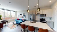 This thoughtfully designed single-floor residence offers a seamless blend of modern style and functional living. The open-concept kitchen and living area serves as the heart of the home, anchored by a sleek island with a white quartz countertop and leather bar seating for three. The kitchen is outfitted with two-tone cabinetry in light gray and white, a white subway tile backsplash, stainless steel appliances including a refrigerator, range, and microwave, and industrial-style pendant lighting overhead. Wood-look flooring runs throughout this area, adding warmth and continuity.

The living space flows naturally from the kitchen, featuring large windows that flood the room with natural light and offer views of the surrounding neighborhood. A vibrant blue sectional sofa and an eclectic gallery wall with framed art and woven decorative accents bring personality to the space.

The bedroom is generously proportioned, with plush carpeting, a large window overlooking the outdoors, and a trio of circular decorative mirrors above the bed. The en-suite bathroom features a long floating vanity with ample storage, a white quartz countertop, a well-lit mirror, and hardwood-style flooring. An in-unit laundry area completes this well-rounded residence.