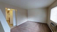 This two-floor space presents a straightforward and functional layout designed to accommodate a range of lifestyles. The pictured room features wall-to-wall carpeting in a warm, neutral tone that adds a layer of comfort underfoot, complemented by smooth, cream-colored walls that create a bright and open atmosphere. A textured ceiling adds subtle visual interest overhead. Clean white baseboards frame the perimeter of the room, contributing to a tidy, well-maintained appearance. A white paneled door is visible toward one side of the room, providing access to an adjoining area, while a glimpse of hard-surface flooring beyond the doorway suggests varied flooring materials throughout the space. Electrical outlets are positioned at standard intervals along the walls, and the overall layout offers a versatile, open floor plan. The neutral color palette throughout creates a blank canvas, making it easy to personalize the space. Spanning two floors, the property provides separation between living and private areas, supporting a functional and organized flow throughout the home.