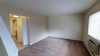 This two-floor space presents a straightforward and functional layout designed to accommodate a range of lifestyles. The pictured room features wall-to-wall carpeting in a warm, neutral tone that adds a layer of comfort underfoot, complemented by smooth, cream-colored walls that create a bright and open atmosphere. A textured ceiling adds subtle visual interest overhead. Clean white baseboards frame the perimeter of the room, contributing to a tidy, well-maintained appearance. A white paneled door is visible toward one side of the room, providing access to an adjoining area, while a glimpse of hard-surface flooring beyond the doorway suggests varied flooring materials throughout the space. Electrical outlets are positioned at standard intervals along the walls, and the overall layout offers a versatile, open floor plan. The neutral color palette throughout creates a blank canvas, making it easy to personalize the space. Spanning two floors, the property provides separation between living and private areas, supporting a functional and organized flow throughout the home.