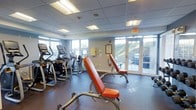This fitness center is a dedicated exercise facility designed to support a well-rounded workout routine. The space is outfitted with a variety of cardio equipment, including elliptical trainers and upright exercise machines, arranged to maximize floor space and ease of movement. Strength training is supported by adjustable weight benches with orange upholstered padding, providing a functional and practical setup for resistance exercises.

The flooring is a durable rubber gym surface, ideal for high-impact activity and equipment placement. Overhead, a drop tile ceiling with recessed fluorescent lighting ensures the space is consistently and evenly illuminated. Large windows along one wall and full-length glass doors flood the interior with natural light while offering views of the surrounding outdoor landscape, including a hillside backdrop that adds a refreshing visual element to workouts.

The layout is open and organized, allowing for comfortable circulation between equipment. Safety features such as a fire extinguisher and clearly marked exit signage are present throughout. Informational postings are displayed on the wall, providing facility guidelines for users. Overall, this fitness center offers a clean, functional, and well-lit environment suited for daily exercise.