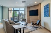 Located at 2725 Marshall Ct in Shorewood Hills, WI, this contemporary community amenity space offers a well-appointed interior complemented by an expansive outdoor terrace. The interior is finished with large-format light tile flooring, soft blue-gray accent walls, and warm wood baseboard trim that together create a calm, modern atmosphere. The lounge area features a comfortable sectional sofa, mid-century-inspired accent chairs, and a marble-top coffee table arranged around a wall-mounted flat-screen television set against a striking horizontal wood-panel feature wall. Vibrant blue abstract artwork adds a curated, gallery-like touch throughout. A dedicated dining area accommodates a large rectangular table with upholstered chairs, illuminated by a series of contemporary cylindrical pendant lights. A built-in kitchenette with cabinetry, a sink, and under-counter refrigeration provides convenient refreshment service, while open floating shelves offer decorative display space. Large glass doors open directly onto the outdoor terrace, which is generously furnished with lounge chairs, dining sets, and shaded umbrella tables amid colorful planters. The terrace offers open views of the surrounding Shorewood Hills streetscape and tree-lined neighborhood, making it an ideal setting for both relaxation and social gatherings.