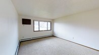 This single-floor space offers a clean, functional layout designed for comfortable everyday living. The main living area is notably open and bright, anchored by a large multi-pane window that allows ample natural light to fill the room, creating an airy and welcoming atmosphere. The window features classic brown trim framing, adding a subtle contrast to the crisp white walls that surround the space. Underfoot, neutral-toned wall-to-wall carpeting runs throughout, providing warmth and a quiet, comfortable feel to the interior. A baseboard heating system lines the perimeter of the living area, ensuring consistent climate control throughout the colder months. The ceiling features a textured finish that adds subtle depth and character to the otherwise clean and minimalist aesthetic. A wall-mounted unit, likely a thermostat or ventilation control, is positioned near the window for easy access. The white walls are clean and unadorned, offering a blank canvas that allows for flexible interior customization. Overall, the space presents a straightforward, well-maintained environment that is both practical and inviting, making it well-suited for a variety of uses.