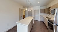 This single-floor space showcases a contemporary design with a cohesive blend of warm and neutral tones throughout. The kitchen serves as a central highlight, featuring light wood-toned cabinetry with open shelving, a classic white subway tile backsplash, stainless steel appliances including a range, microwave, and dishwasher, and a spacious island with a light quartz countertop illuminated by pendant lighting above. Dark hardwood-style flooring runs through the kitchen and living areas, adding warmth and contrast to the bright, neutral walls.

The bedroom is a comfortable, carpeted retreat filled with natural light through wide horizontal blinds, complemented by a ceiling fan that enhances airflow and comfort. The room's clean, unadorned walls offer flexibility for personalization.

The bathroom is finished with a full soaking tub and shower combination surrounded by white subway tile, a built-in niche for storage, and an adjacent open shelving unit for linens. Wood-look flooring continues here, paired with a modern floating-style vanity. Together, the rooms create a cohesive, functional living environment with clean modern finishes throughout.