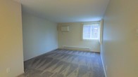 This single-floor space features a generously sized main room finished with patterned wall-to-wall carpeting in neutral grey tones, providing a practical and comfortable foundation. The walls are painted in a soft, warm beige, contributing to a clean and understated aesthetic throughout the room. A wall-mounted air conditioning unit is positioned on one wall, offering convenient climate control, while a baseboard heating unit runs along the base of the exterior wall, ensuring efficient year-round temperature regulation. Natural light enters the room through a single window fitted with white vertical blinds, allowing for adjustable light control and privacy. The window also features a white security grille, adding an element of safety. The ceiling is smooth and standard height, keeping the space feeling open and easy to maintain. Overall, the room presents a straightforward, functional layout well-suited for a variety of uses, with essential utilities already in place and a neutral palette that accommodates diverse furnishing and décor choices.