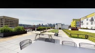 This expansive rooftop terrace offers a well-appointed outdoor retreat with striking panoramic views of a downtown city skyline. The space is designed for both relaxation and active recreation, featuring a contemporary concrete ping-pong table as a central focal point, surrounded by modern wire-frame chairs that provide casual seating. A generous patch of manicured green lawn occupies one side of the terrace, offering a refreshing contrast to the clean, large-format tile paving that covers the main gathering area. Raised planter beds with lush greenery line portions of the perimeter, adding natural texture and softness to the hardscape. Additional seating is available throughout, including picnic-style tables positioned toward the edges, encouraging residents or visitors to take in the open skyline views. The rooftop's open layout allows for flexible use, accommodating group gatherings or quiet outdoor lounging. The surrounding urban backdrop, featuring cranes, neighboring buildings, and the distant city skyline, gives the terrace a distinctly metropolitan character, while the open sky above ensures an abundance of natural light throughout the day.