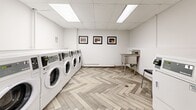 Located at 1255 Pendrell Street in Vancouver's West End, this shared laundry facility offers a clean, well-maintained, and efficiently organized space designed for everyday use. The room accommodates multiple front-loading washing machines and dryers arranged along one wall, providing ample capacity for residents. A utility sink station offers added convenience for hand-washing or pre-treating garments, while a stainless steel folding table with a lower shelf provides a dedicated surface for sorting and folding laundry. The floor features a striking herringbone-patterned vinyl plank flooring in soft, neutral tones that adds a refined visual element to an otherwise functional space. The ceiling is fitted with a drop tile grid and recessed fluorescent light panels, ensuring bright, even illumination throughout. White cabinetry beneath an information board offers additional storage, and a cork bulletin board displays notices and instructions for users. The walls are finished in crisp white, contributing to the room's clean and welcoming ambiance. A wall-mounted clock and clearly posted guidelines keep the space orderly and user-friendly. A fire-rated entry door with a glazed panel provides secure, easy access. Overall, the facility combines practicality with thoughtful design details to deliver a comfortable and functional laundry experience.
