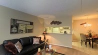 This two-story residence offers a thoughtfully arranged interior highlighted by an open-concept design that visually connects the living room and kitchen. The living area is carpeted and features a textured ceiling, with warm, neutral-toned walls that create an inviting atmosphere. A black leather sofa serves as the focal point of the living space, complemented by a glass-top coffee table and decorative accents with a distinct movie theater theme, including framed film-inspired wall art and decorative film reel wall pieces. A table lamp provides warm ambient lighting to the space. A half-wall pass-through connects the living room to the kitchen, offering both visual openness and a sense of defined space. The kitchen is visible through this opening and features white cabinetry and appliances, including a refrigerator, with additional storage and counter space visible from the living area. A hallway extends beyond the living space, suggesting further rooms and access to the second floor. Overall, the layout balances functionality with a personalized, character-filled aesthetic throughout the main level.