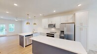 Located at 2312 Ben Craig Dr in Charlotte, NC, this contemporary 4-story townhome offers 3 bedrooms and 5 bathrooms across a well-designed, generously proportioned floor plan. The entry level features an attached two-car garage with drywall-finished walls, concrete flooring, and interior stair access leading up into the main living areas. The second floor hosts an open-concept kitchen and living room bathed in natural light from multiple windows. The kitchen is outfitted with white shaker-style cabinetry, quartz countertops, stainless steel appliances including a French door refrigerator and range with microwave, and a contrasting navy island — all complemented by light hardwood-style flooring throughout. The bathrooms are clean and bright, featuring white fixtures, tub-shower combinations, and a primary bath with a dual-sink vanity topped with quartz counters and a walk-in shower. Bedrooms are spread across the upper floors, with light hardwood flooring in the main bedrooms and plush carpeting in the top-floor room, which features vaulted ceilings. Each bedroom is bright and airy, with ceiling fans and natural light. The exterior showcases a classic white siding facade set within a well-maintained, newly developed neighborhood community in Charlotte.