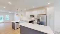 Located at 2312 Ben Craig Dr in Charlotte, NC, this contemporary 4-story townhome offers 3 bedrooms and 5 bathrooms across a well-designed, generously proportioned floor plan. The entry level features an attached two-car garage with drywall-finished walls, concrete flooring, and interior stair access leading up into the main living areas. The second floor hosts an open-concept kitchen and living room bathed in natural light from multiple windows. The kitchen is outfitted with white shaker-style cabinetry, quartz countertops, stainless steel appliances including a French door refrigerator and range with microwave, and a contrasting navy island — all complemented by light hardwood-style flooring throughout. The bathrooms are clean and bright, featuring white fixtures, tub-shower combinations, and a primary bath with a dual-sink vanity topped with quartz counters and a walk-in shower. Bedrooms are spread across the upper floors, with light hardwood flooring in the main bedrooms and plush carpeting in the top-floor room, which features vaulted ceilings. Each bedroom is bright and airy, with ceiling fans and natural light. The exterior showcases a classic white siding facade set within a well-maintained, newly developed neighborhood community in Charlotte.