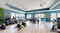 Located at 111 Teal Cir in Little River, SC, this fitness facility offers a comprehensive, single-floor layout designed to accommodate a wide range of workout activities. The main exercise room is a spacious, vibrant area painted in a bold teal-blue palette and finished with light wood-look flooring throughout. It is outfitted with an extensive array of equipment, including treadmills, ellipticals, stationary bikes, rowing machines, and a variety of strength training machines, as well as a full dumbbell rack. Ceiling fans and recessed lighting ensure a comfortable and well-lit environment. A striking green living wall accent adds a fresh, energizing element to the space, while wall-mounted mirrors enhance the sense of openness. A dedicated studio room, bathed in abundant natural light through large windows and clerestory panes, features floor-to-ceiling mirrors and light wood-look flooring, making it ideal for yoga, stretching, or group classes. Views of the surrounding community, including a pool area, can be seen through the studio windows. The facility is connected by a series of hallways with soft blue walls and built-in shelving, providing practical transitional spaces and easy navigation throughout.
