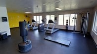 This single-floor fitness room offers a well-equipped workout environment designed to support a variety of exercise routines. The space features durable dark rubber flooring throughout, providing a stable and impact-resistant surface ideal for both cardio and strength training activities. A bold yellow accent wall adds an energetic visual element, complementing the otherwise neutral color palette of white walls and a textured ceiling fitted with fluorescent lighting panels.

The room is lined with several windows along one wall, allowing generous natural light to fill the space and offering views of the surrounding outdoor greenery. Cardio equipment is a primary focus, with multiple treadmills and an elliptical or stair climber machine positioned to take advantage of the natural light. A freestanding heavy punching bag anchors one area of the room, ideal for boxing or functional training. A rack of weighted plates and a cable resistance station provide strength training options, while exercise mats laid out on the floor offer space for stretching or floor-based workouts. A wall-mounted fan enhances air circulation throughout the room. Overall, the space delivers a functional and motivating fitness environment.