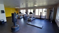 This single-floor fitness room offers a well-equipped workout environment designed to support a variety of exercise routines. The space features durable dark rubber flooring throughout, providing a stable and impact-resistant surface ideal for both cardio and strength training activities. A bold yellow accent wall adds an energetic visual element, complementing the otherwise neutral color palette of white walls and a textured ceiling fitted with fluorescent lighting panels.

The room is lined with several windows along one wall, allowing generous natural light to fill the space and offering views of the surrounding outdoor greenery. Cardio equipment is a primary focus, with multiple treadmills and an elliptical or stair climber machine positioned to take advantage of the natural light. A freestanding heavy punching bag anchors one area of the room, ideal for boxing or functional training. A rack of weighted plates and a cable resistance station provide strength training options, while exercise mats laid out on the floor offer space for stretching or floor-based workouts. A wall-mounted fan enhances air circulation throughout the room. Overall, the space delivers a functional and motivating fitness environment.