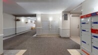Located at 55 Rue Hasting in Dollard-des-Ormeaux, Quebec, this building lobby serves as the primary common entry area for residents. The space is configured in an L-shaped layout, offering a generous and well-organized circulation area finished with a combination of tile flooring and wall-to-wall carpeting throughout the main corridor.

The lobby's design is clean and functional, with neutral beige walls accented by a continuous white chair rail that runs the length of the hallways. Decorative wall-mounted planters add a welcoming touch of greenery along the corridor. Recessed and wall-mounted lighting ensures the space is consistently well-lit throughout.

A key feature of the lobby is the dedicated mail area, which houses a large bank of individual resident mailboxes along one wall, alongside a Canada Post parcel locker unit for secure package delivery. An elevator provides convenient vertical access to the building's upper floors, complemented by a secured stairwell door equipped with emergency exit signage.

The main corridor leads directly to the building's front entrance, framed by glass double doors that allow natural light to filter in, creating a bright and inviting atmosphere at the entryway. Wooden bench seating and a small wooden planter box are thoughtfully placed throughout, adding warmth and practicality to this well-appointed residential lobby.