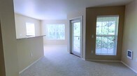 This single-floor residence offers a welcoming and well-lit living environment designed for comfortable everyday living. The main living area is generously sized, finished with soft wall-to-wall carpeting that adds warmth underfoot. Neutral, light-toned walls complement the airy atmosphere throughout the space.

Natural light is a defining feature of this residence, with multiple large windows fitted with horizontal blinds allowing for adjustable brightness. A sliding glass door leads directly to an outdoor balcony, offering pleasant views of mature trees and lush greenery — bringing a sense of the outdoors inside.

A partial wall separates the living area from what appears to be a kitchen or dining counter space, providing an open, connected layout that supports easy flow between living and dining functions. The clean lines and simple finishes of the interior create a neutral canvas well-suited for a variety of furnishing styles.

Overall, the space presents a bright, calm, and functional atmosphere, with an emphasis on natural light and outdoor connectivity. The greenery visible through the windows and balcony door adds a refreshing natural element that enhances the overall ambiance of the home.