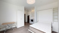 This modern studio unit is thoughtfully designed to maximize functionality within a compact footprint. The open-concept layout seamlessly integrates the sleeping area, kitchenette, and living space into a single cohesive environment. The interior is finished in a crisp, all-white palette, with smooth ceilings, recessed lighting, and a flush overhead light fixture that collectively create a bright and airy ambiance. Durable wood-look laminate flooring runs throughout, adding warmth and visual continuity to the space. The sleeping area features a Murphy-style wall bed, offering flexibility and freeing up floor space when not in use. A tall, two-door wardrobe provides generous storage, while a compact desk and chair offer a dedicated workspace. The kitchenette is neatly tucked into an alcove and equipped with light grey cabinetry, a stainless steel refrigerator, a built-in microwave, and a small sink — providing all the essential conveniences in an efficient arrangement. A recessed entryway leads to what appears to be a bathroom, completing the unit's practical layout. Clean lines, neutral tones, and smart space-saving solutions define the overall character of this well-appointed studio.