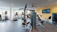 Located at 101 E Main St in downtown Rock Hill, South Carolina, this fitness center is a comprehensively outfitted exercise facility designed to accommodate a wide range of workout routines. The space is laid out on a single floor and is divided into distinct functional zones, each tailored to a specific type of training.

The cardio area features multiple treadmills and elliptical machines, positioned near large windows that allow natural light to fill the space and provide outdoor views. A ceiling fan overhead aids in air circulation during workouts. The flooring throughout is a durable speckled rubber-style surface, well-suited for high-traffic exercise use.

The strength training section is anchored by a Matrix cable and multi-functional machine, complemented by an adjustable bench and additional resistance equipment. A full wall of mirrors expands the visual depth of the room while allowing users to monitor their form. A mounted flat-screen television adds to the amenity offerings.

The free weight area includes a well-stocked dumbbell rack with a broad range of weights, stability balls, and a step platform. A separate functional training zone offers a multi-purpose rig equipped with TRX suspension straps and a heavy punching bag, set against a warm yellow accent wall. Recessed lighting throughout ensures consistent illumination across all areas.